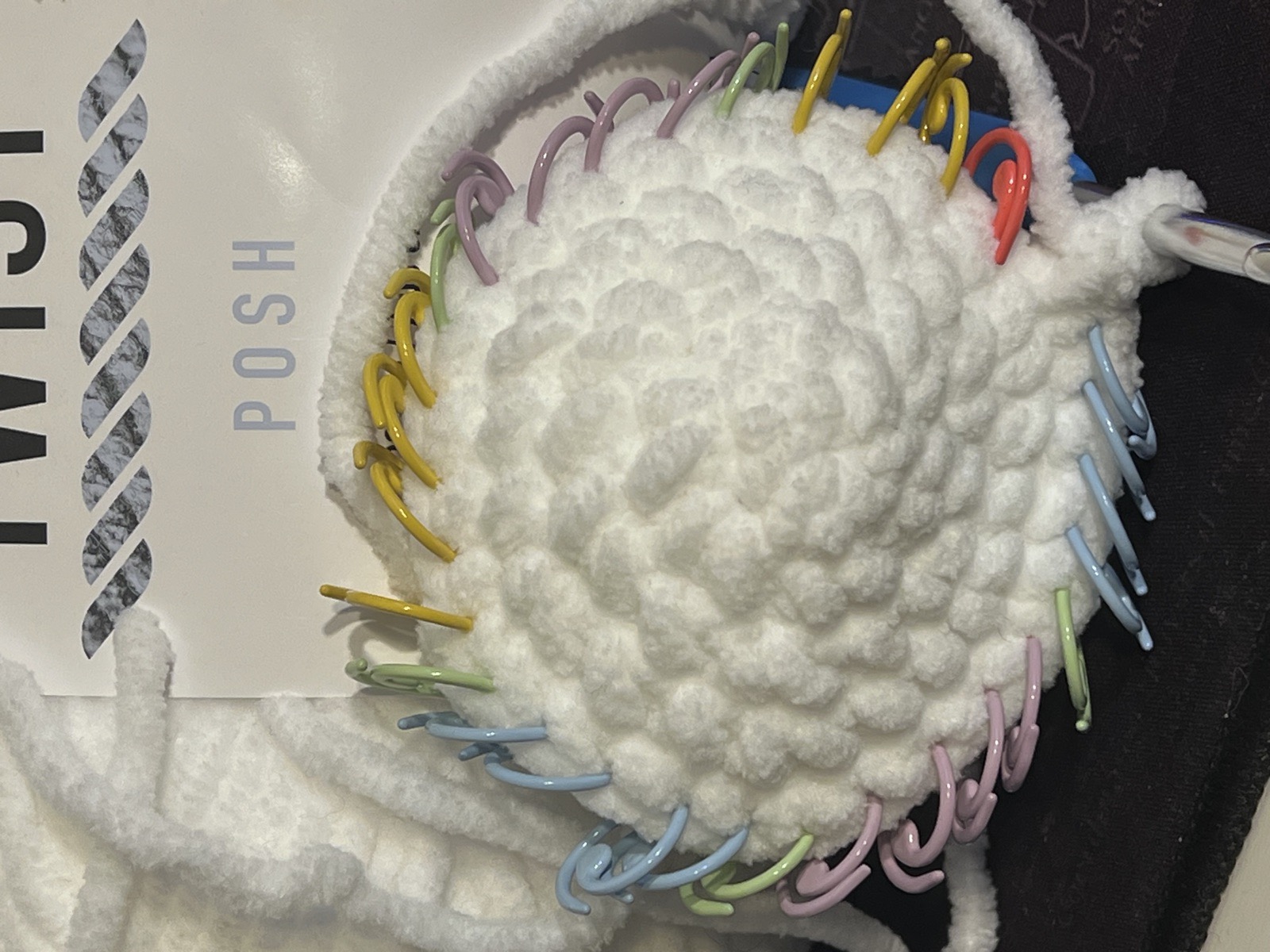 A white amigurumi piece in progress, its edge ringed with pastel stitch markers.