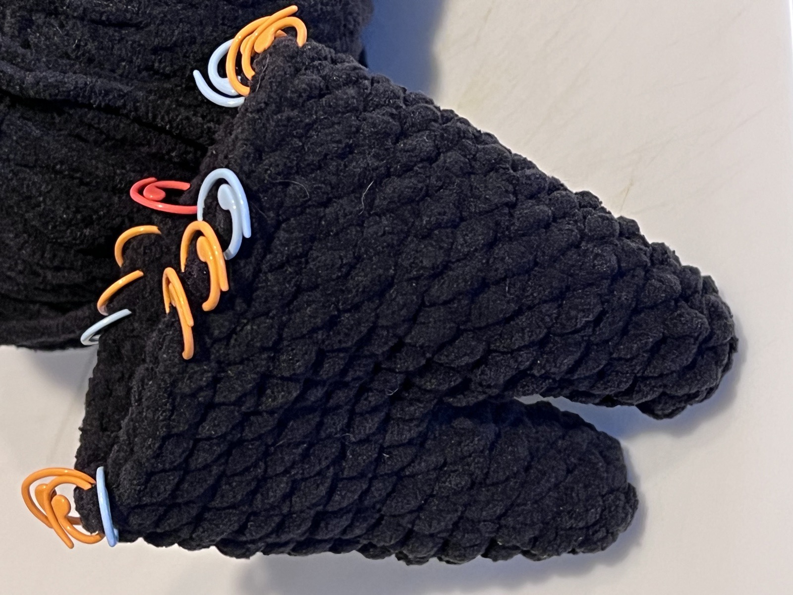Two black amigurumi limbs in progress, tops studded with orange and blue stitch markers.